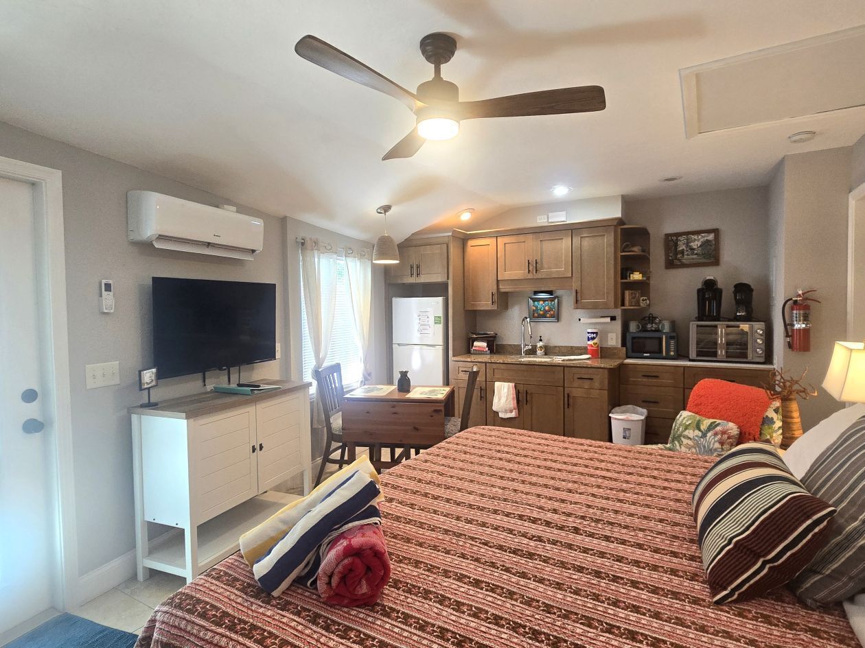 A compact room with a bed, a desk, a television, a kitchenette, and a ceiling fan.