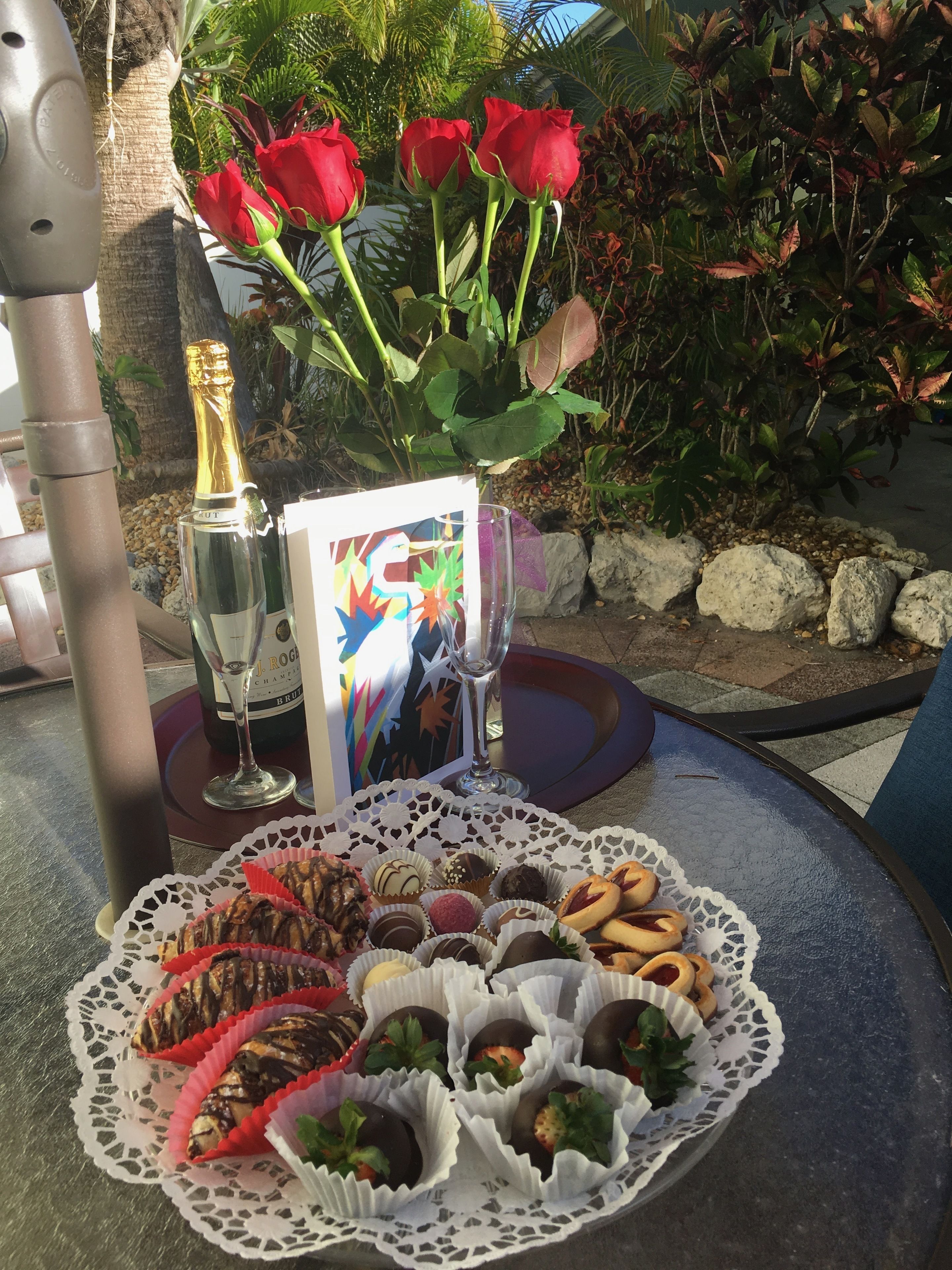 Uma mesa com uma garrafa de champanhe, uma taça, um cartão colorido e um prato com doces variados.