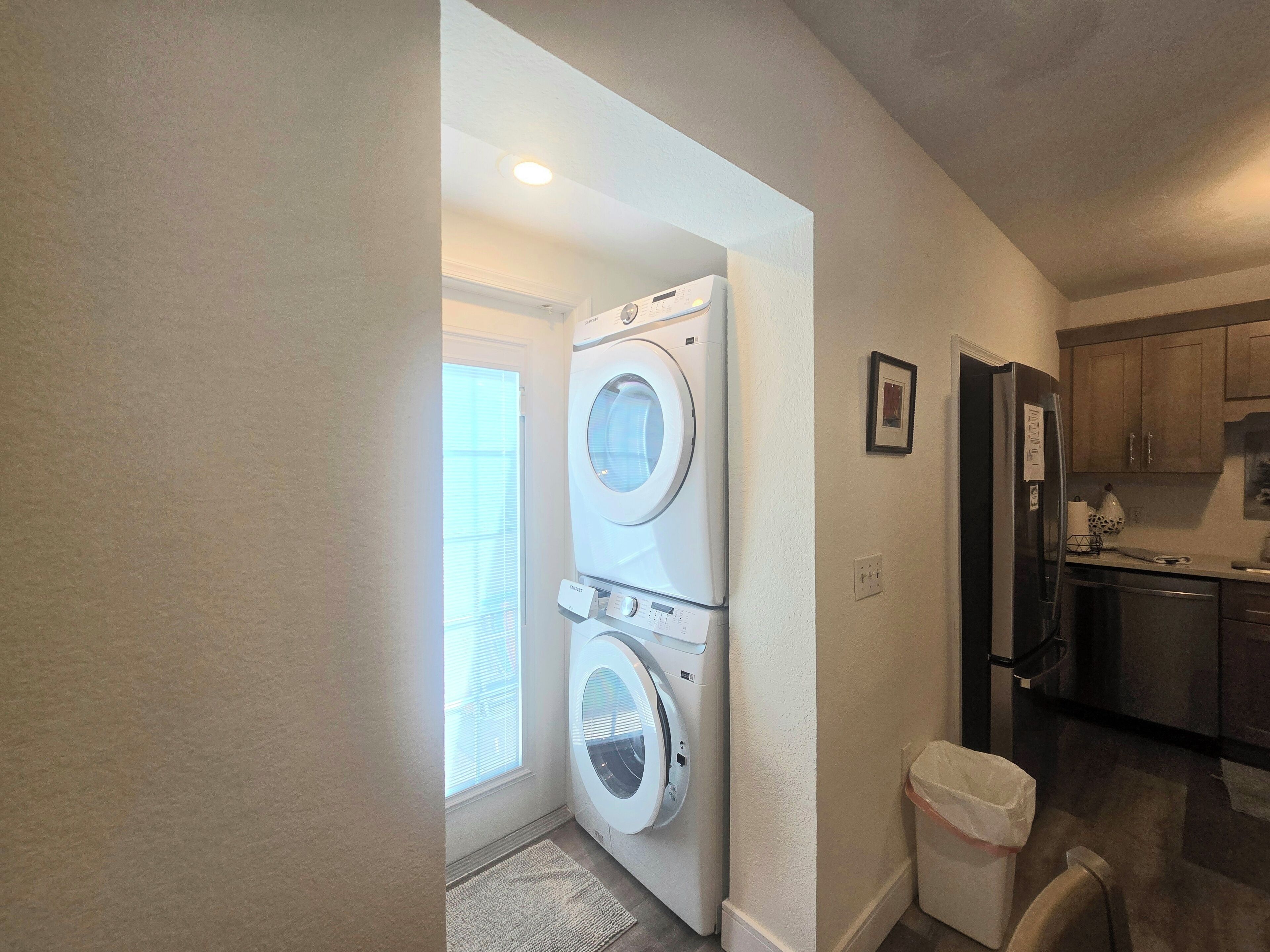 A laundry room with two stacked front-loading washers and dryers, a small trash bin, and a kitchen area with wooden cabinets and a refrigerator.