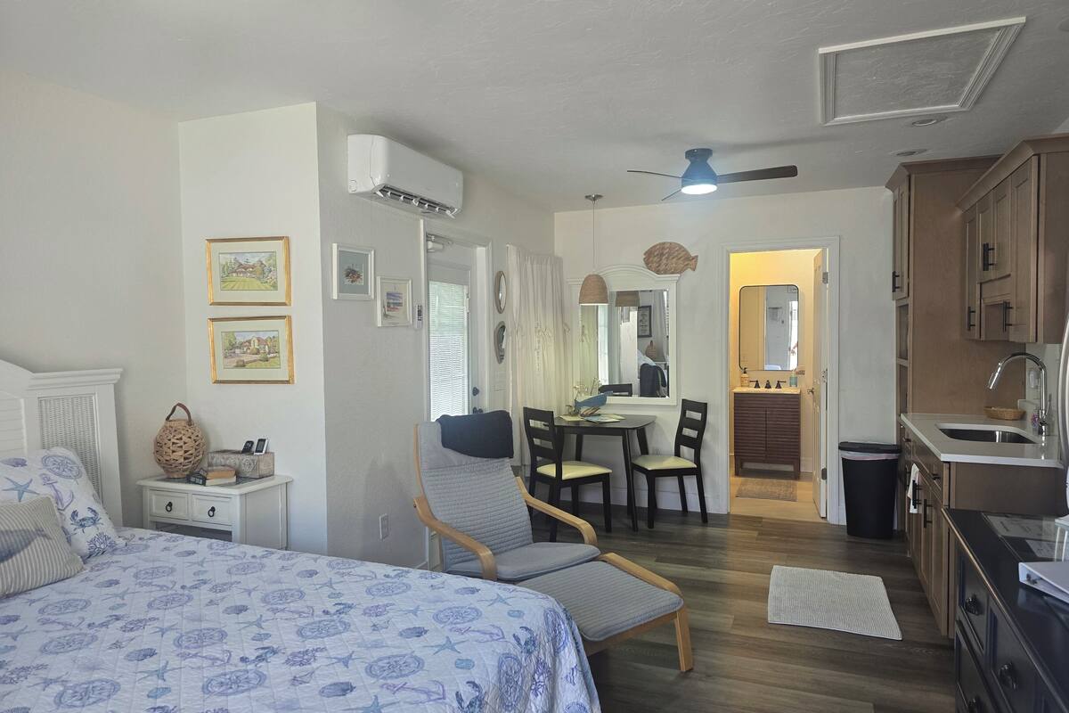 A well-lit, clean room with a bed, a dining table with chairs, a kitchenette, and a ceiling fan.