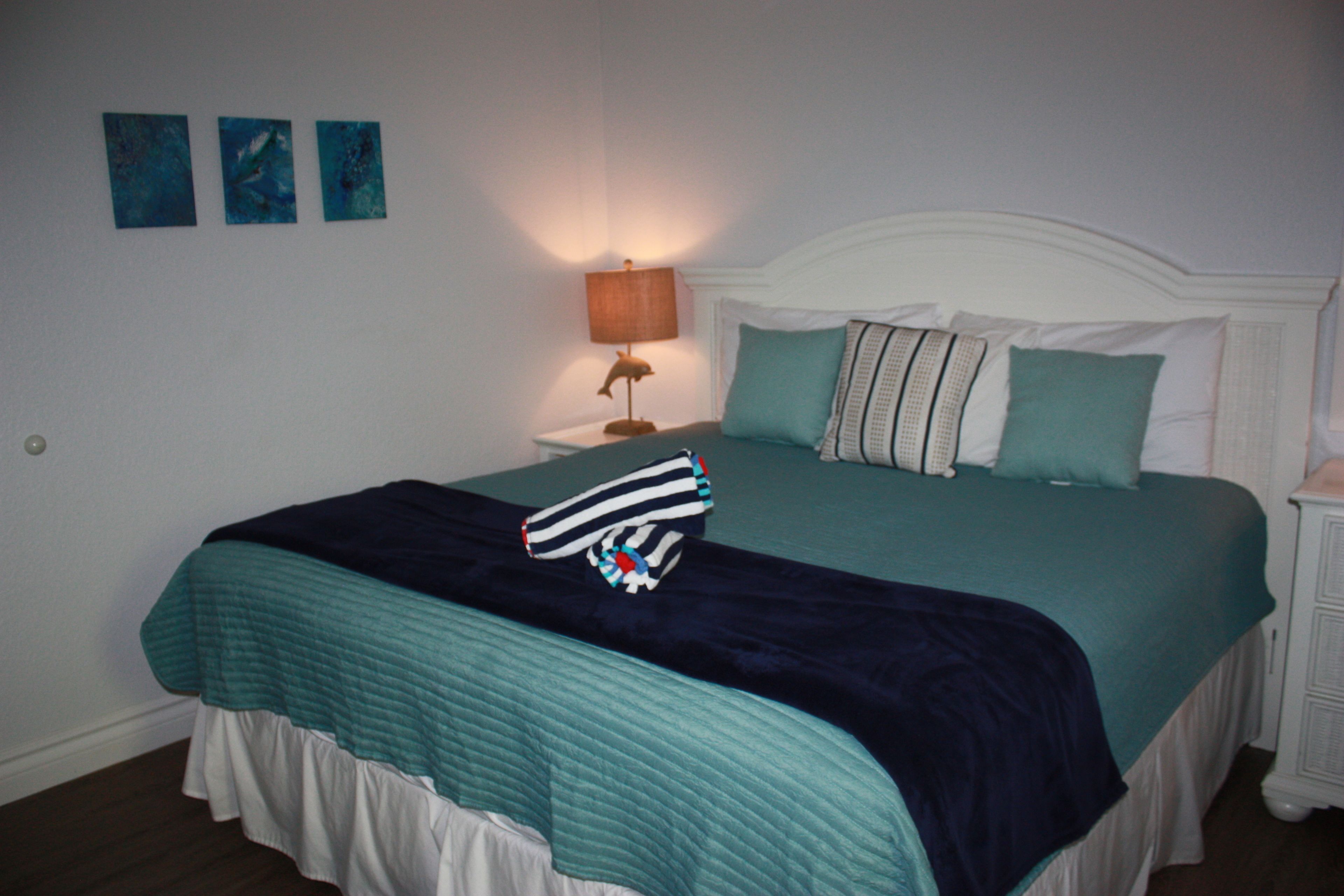 Una cama bien hecha con una colcha verde azulada y ropa de cama blanca, una lámpara de noche y tres cuadros colgados en la pared.