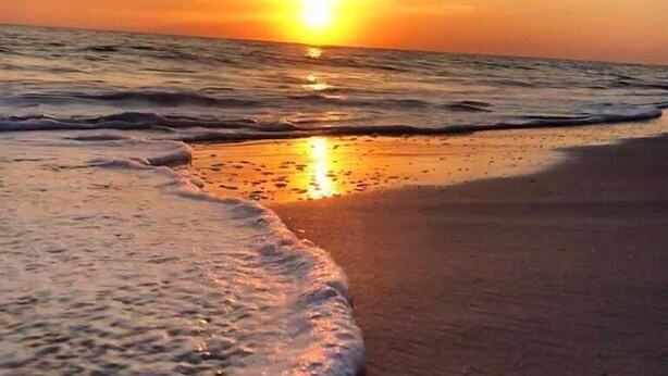 Escena de playa con olas suaves lamiendo la orilla y el sol poniéndose en la distancia.