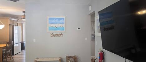 Una sala de estar con un televisor de pantalla plana grande, un cuadro enmarcados de una playa, un banco y una chimenea.