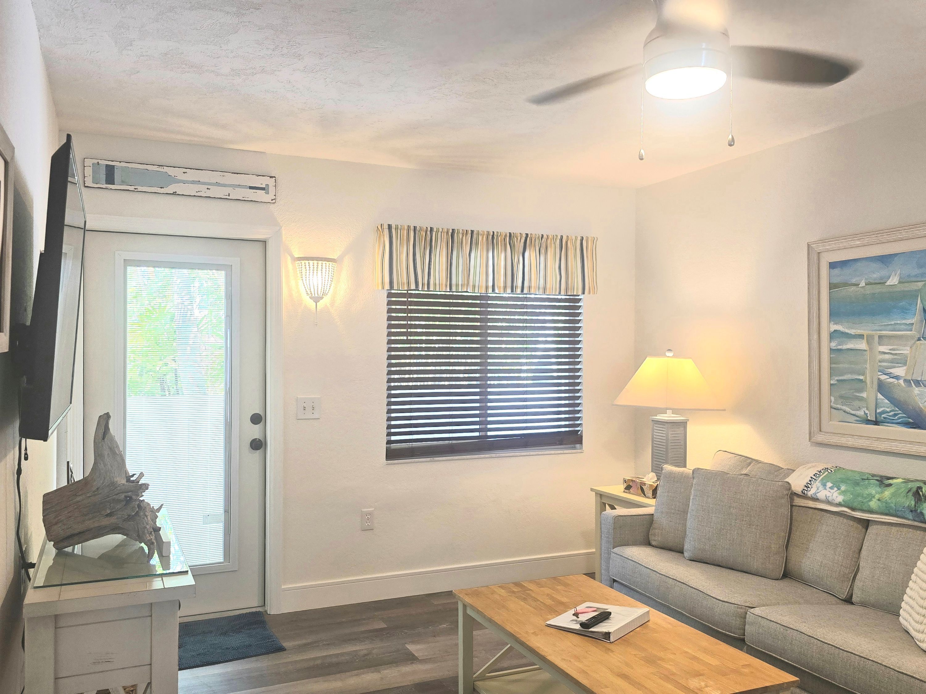 A living room with a sofa, a coffee table, a lamp, a painting, a window with blinds, a ceiling fan, and a door with an air conditioning unit.
