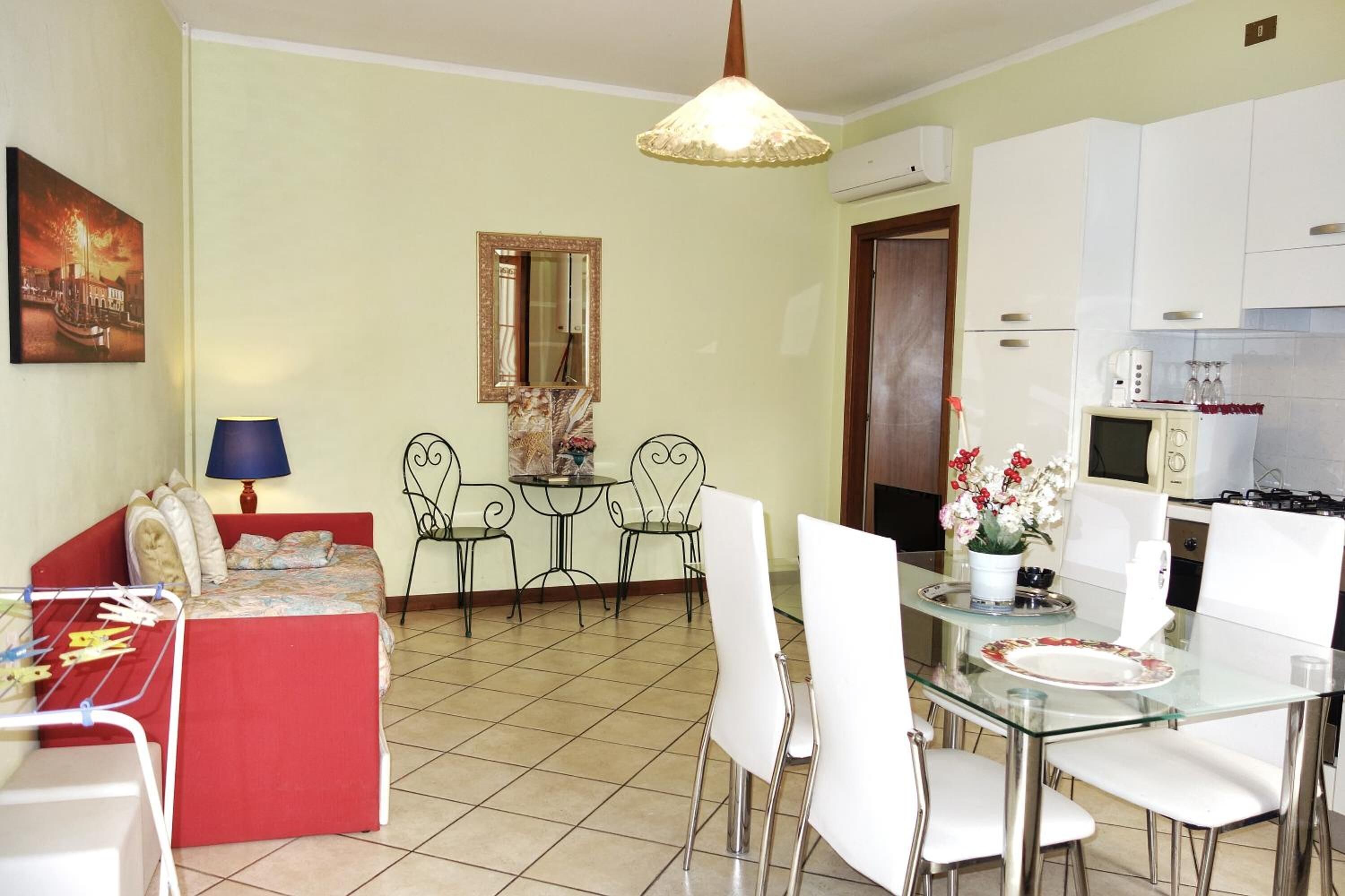 A compact living space with a kitchenette, a dining area with a glass table, a sofa, and a small side table with a lamp.