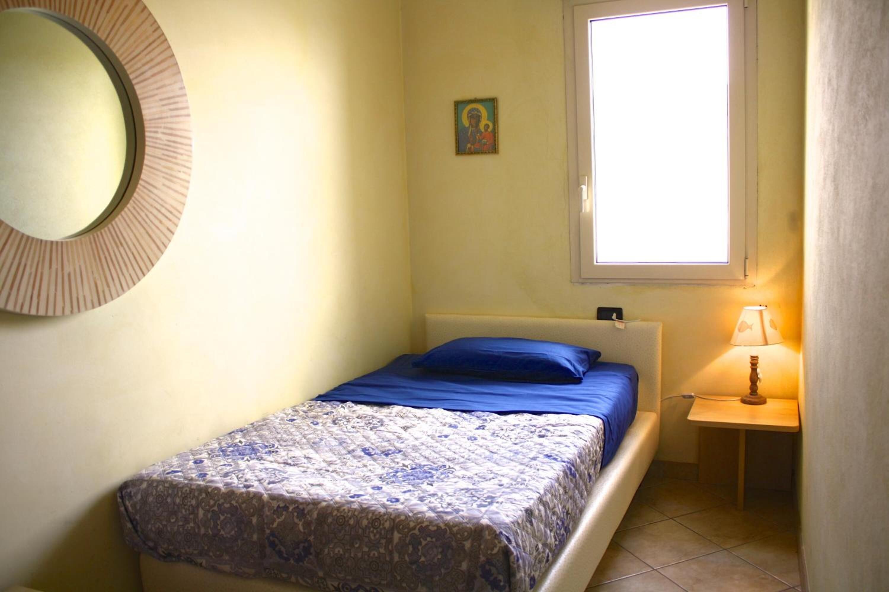 Una cama pequeña y bien hecha con un almohadón azul y una colcha estampada, una mesita de noche con una lámpara, un adorno en la pared y una ventana con luz natural.