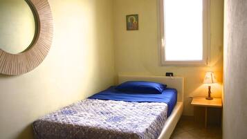 Una cama pequeña y bien hecha con un almohadón azul y una colcha estampada, una mesita de noche con una lámpara, un adorno en la pared y una ventana con luz natural.