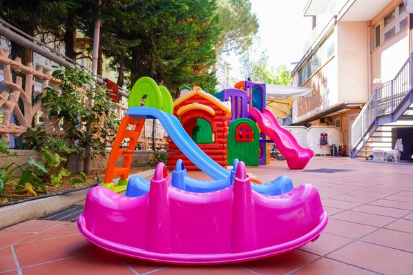 A playground with various colorful play structures, including slides and a climbing ladder, situated in an outdoor area with a tiled floor and surrounded by trees and buildings.