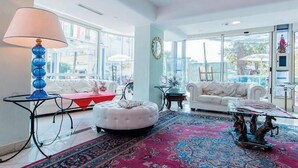 A spacious living room with a large window, white sofas, a red ottoman, and a blue lamp. - Hotel Tridentum (Cesenatico)