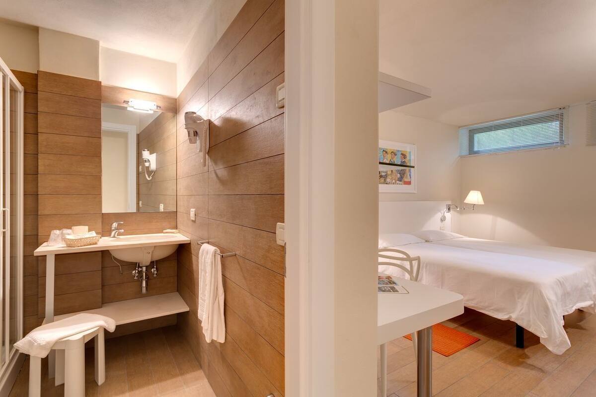 Un baño moderno con pared de madera, un lavamanos y un banco. Se ve una cama con colcha blanca y una mesita de noche con una lámpara.