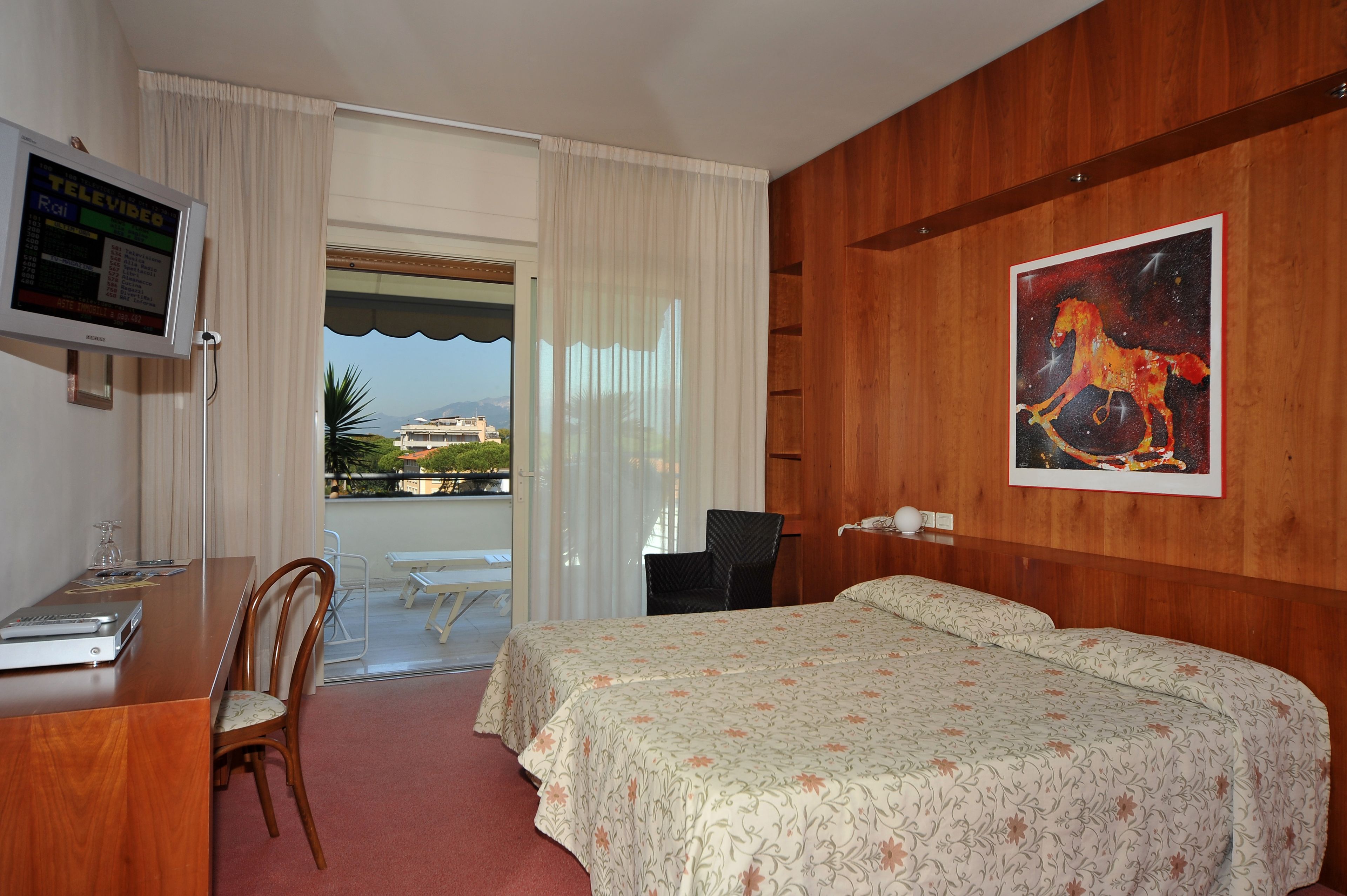 Hotelkamer met een houten wand, een bed met bloemenbeddengoed, een bureau met een televisie en uitzicht op een balkon met tuinmeubilair.
