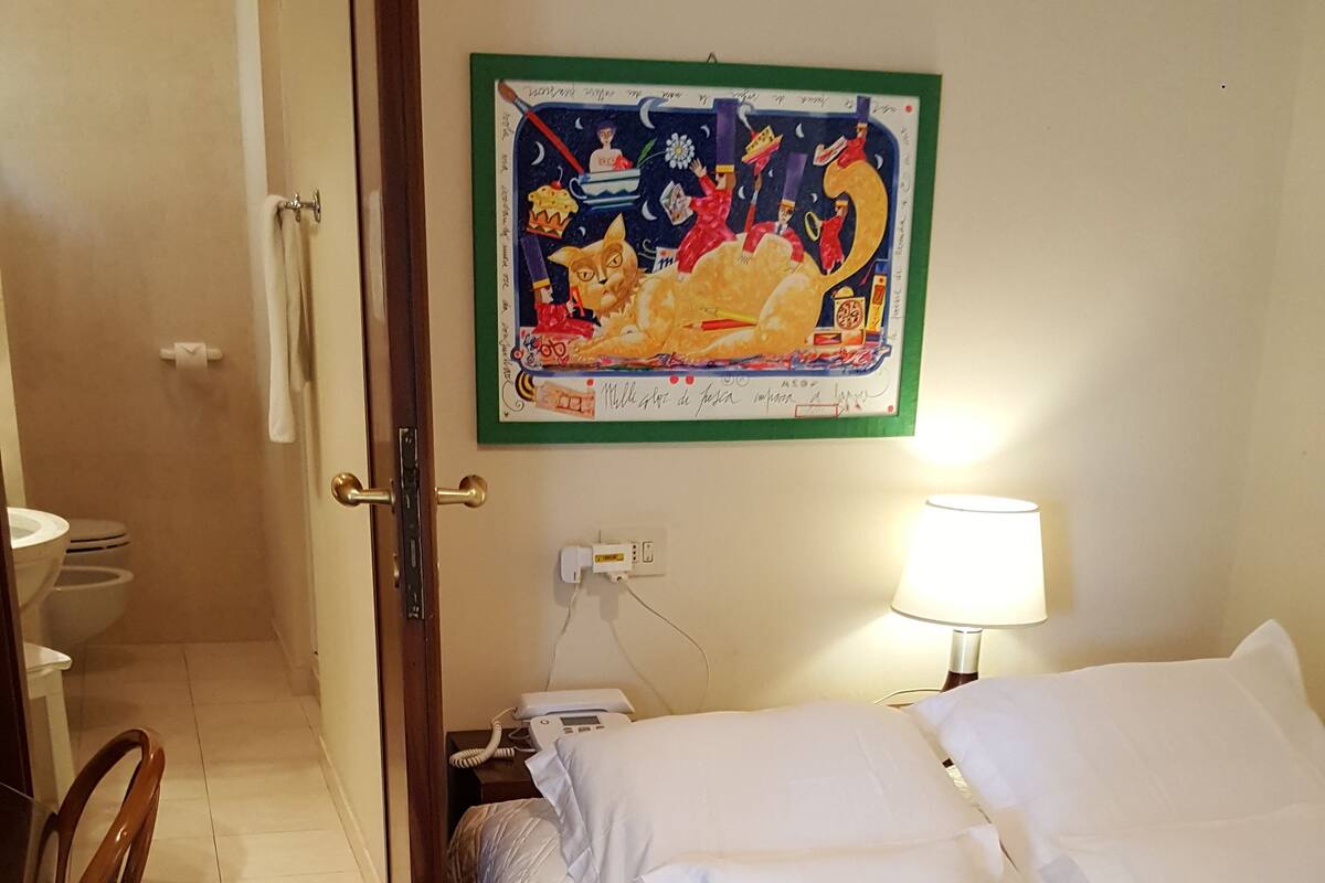 Habitación de hotel con cama, almohadas blancas, mesita de noche con lámpara, cuadro colgado en la pared, baño con inodoro y ducha, y alfombra estampada.