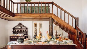 Une pièce spacieuse avec un haut plafond en bois et un escalier menant à un niveau supérieur. Une table à manger est dressée avec différents plats et une statue de coq décorative.