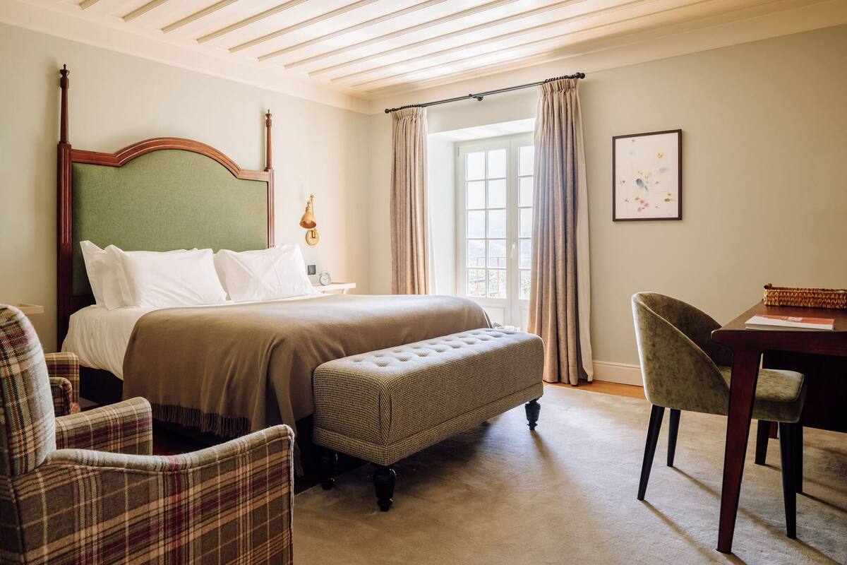 Une chambre d’hôtel avec un grand lit, une banquette, une chaise, un bureau et une fenêtre avec des rideaux.