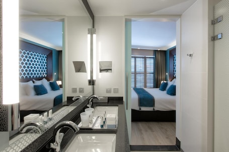 Habitación de hotel moderna con un espejo grande, dos camas, un baño con lavabo y espejo, y vistas al exterior a través de la ventana.