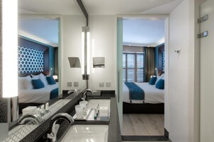 Habitación de hotel moderna con un espejo grande, dos camas, un baño con lavabo y espejo, y vistas al exterior a través de la ventana.