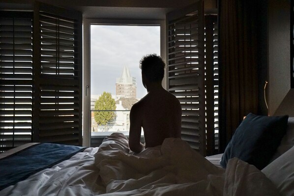 A room with a bed, a window with blinds, and a view of a building outside.