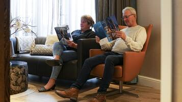 Two people seated on a sofa and a chair, reading books in a living room with a decorative plant and a round side table.