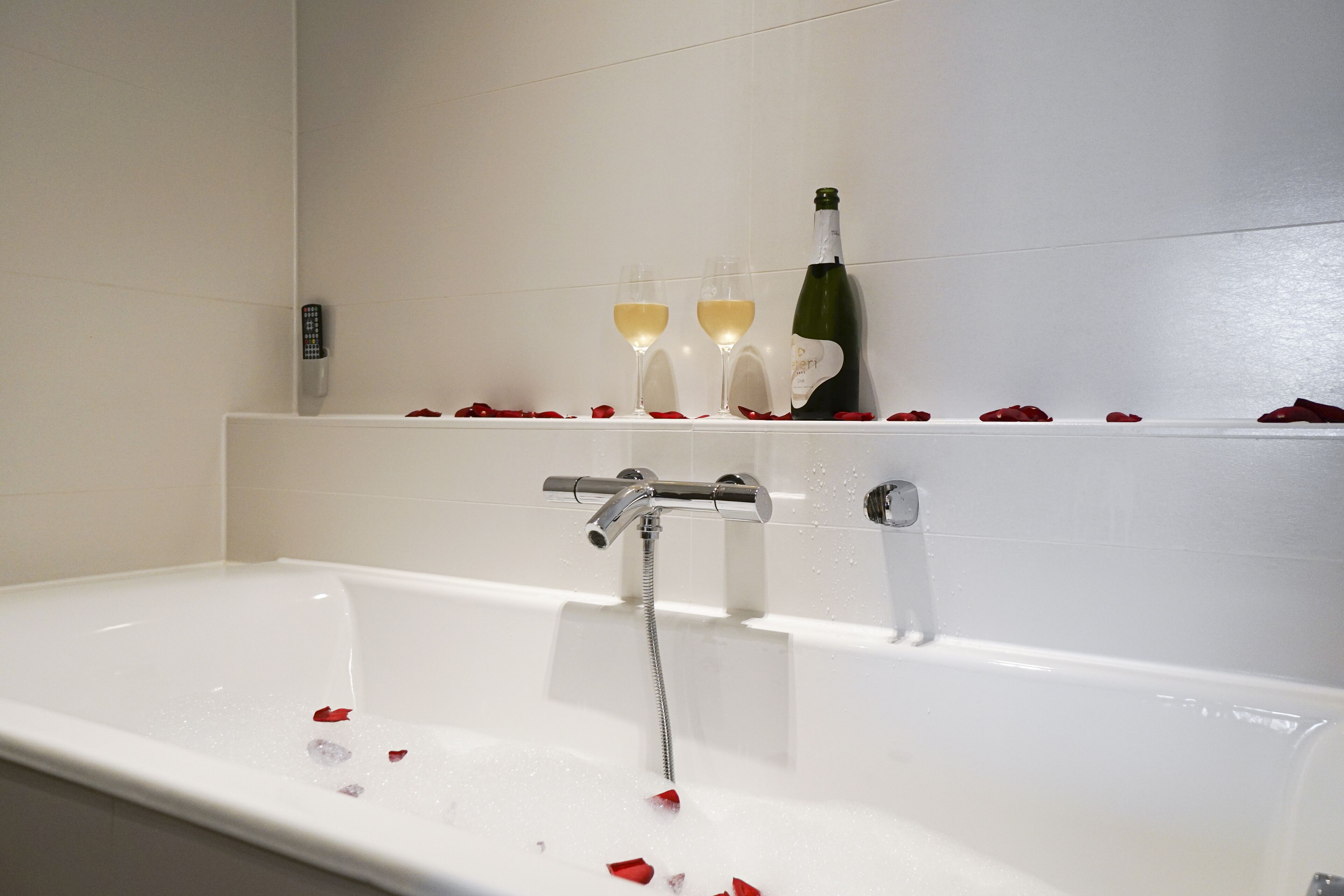 A bathtub with rose petals and champagne glasses on a shelf.
