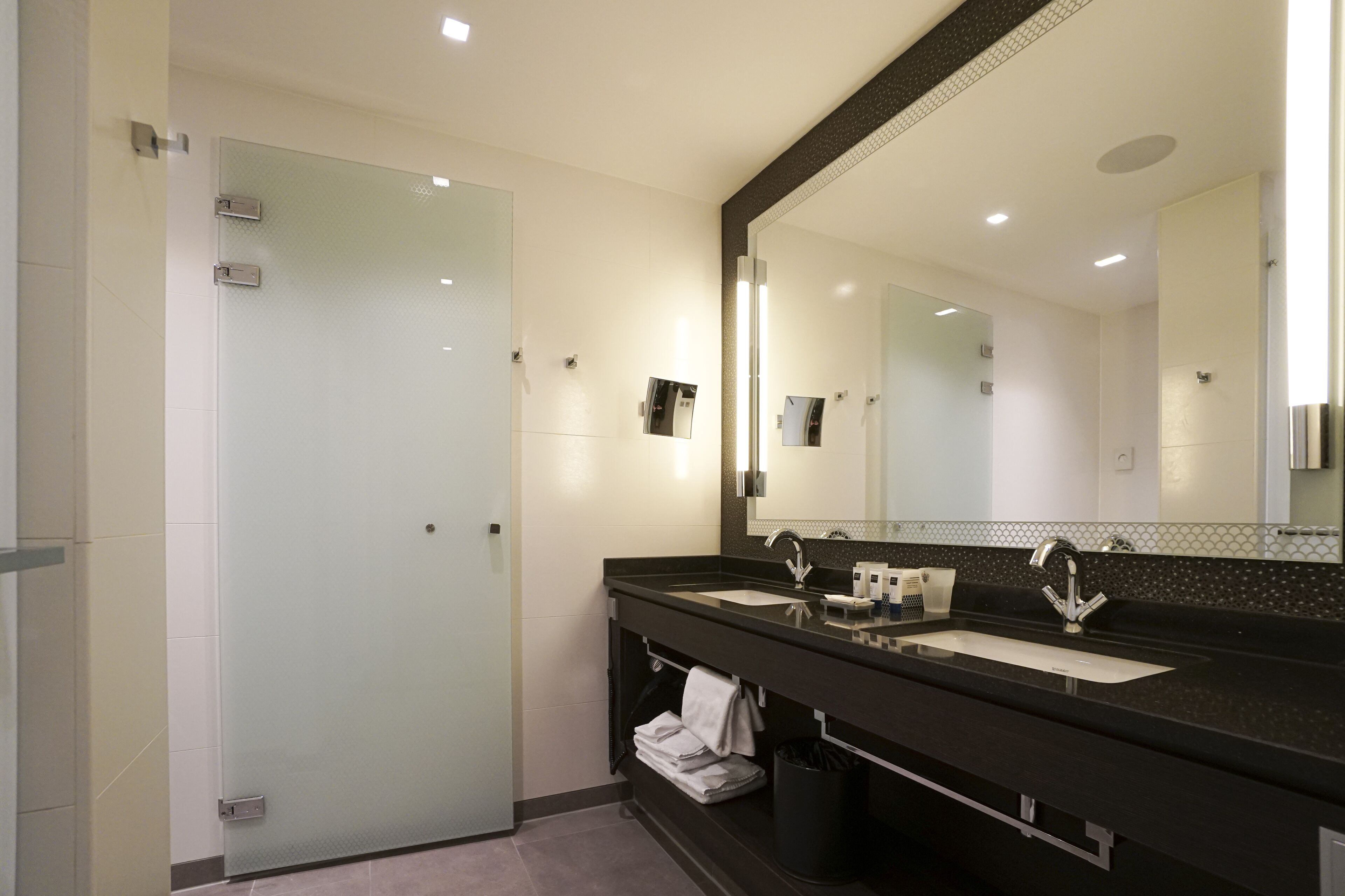 A modern bathroom with a glass shower door, a large mirror, and a black countertop with two sinks.