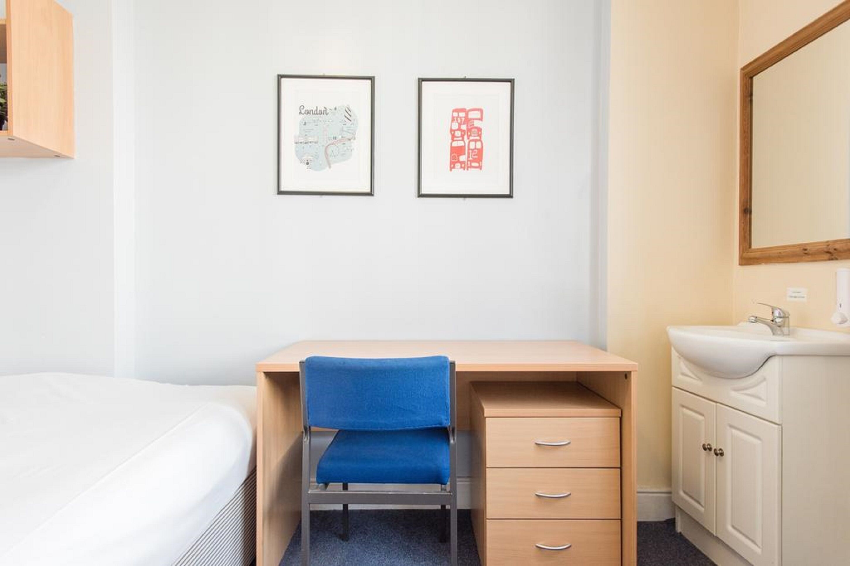 A hotel room with a bed, a desk with a chair, a dresser, a mirror, and two framed pictures on the wall.