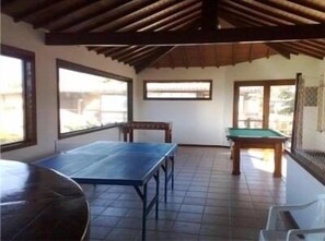 A room with a wooden ceiling, a pool table, a billiards table, and large windows.