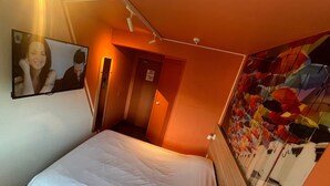 Una camera d'albergo con un letto, una TV a muro, una porta e un murale colorato.