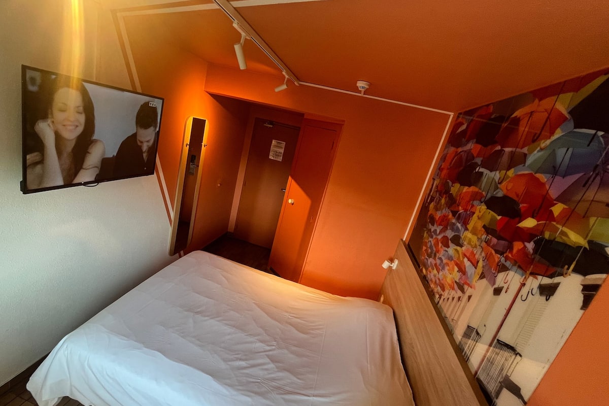Quarto de hotel com uma cama, televisão fixada na parede, uma porta e um mural colorido.