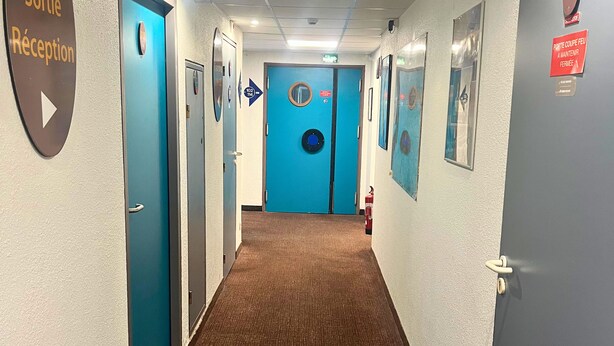 Corridoio di un hotel con porte blu, una segnaletica della reception e frecce direzionali.