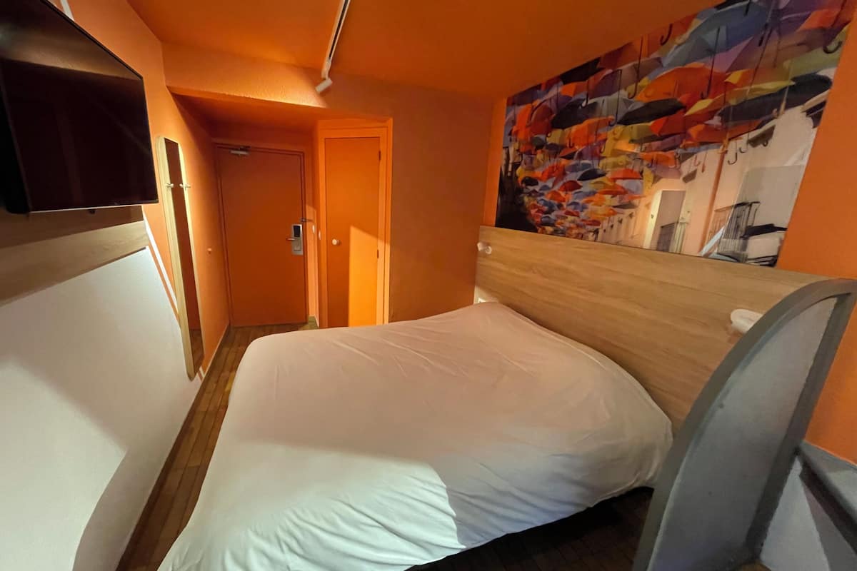 Quarto de hotel com uma cama, televisão fixada na parede e um quadro abstrato colorido.