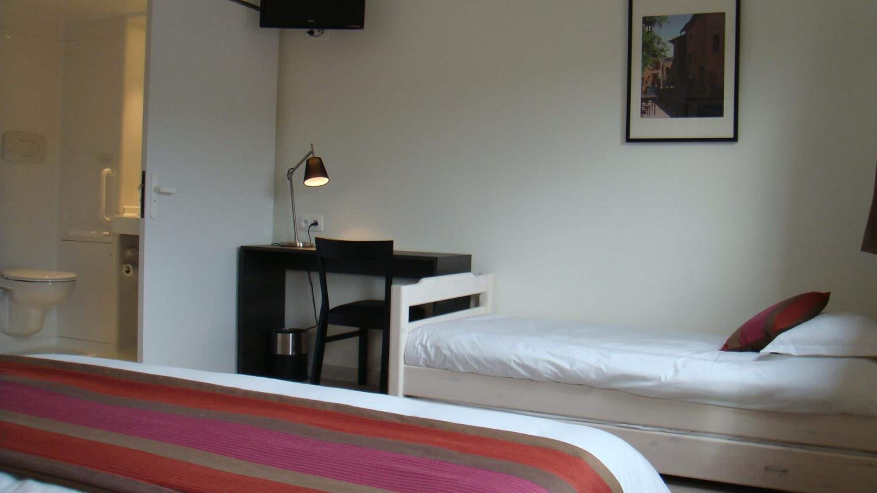 A bedroom with a bed, a desk with a chair, a toilet, and a framed picture on the wall.