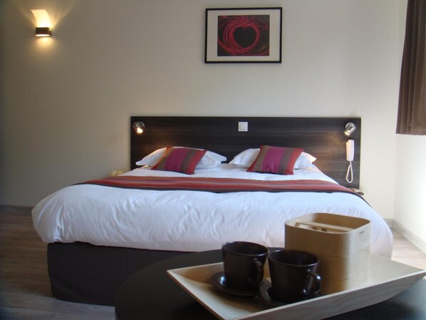 Una cama bien hecha con sábanas blancas y almohadas coloridas, un teléfono en la mesita de noche y una bandeja con dos tazas negras.