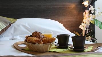 A neatly arranged breakfast tray with croissants, a glass of orange juice, and two black cups on a wooden tray, placed on a bed with white pillows and a dark headboard.