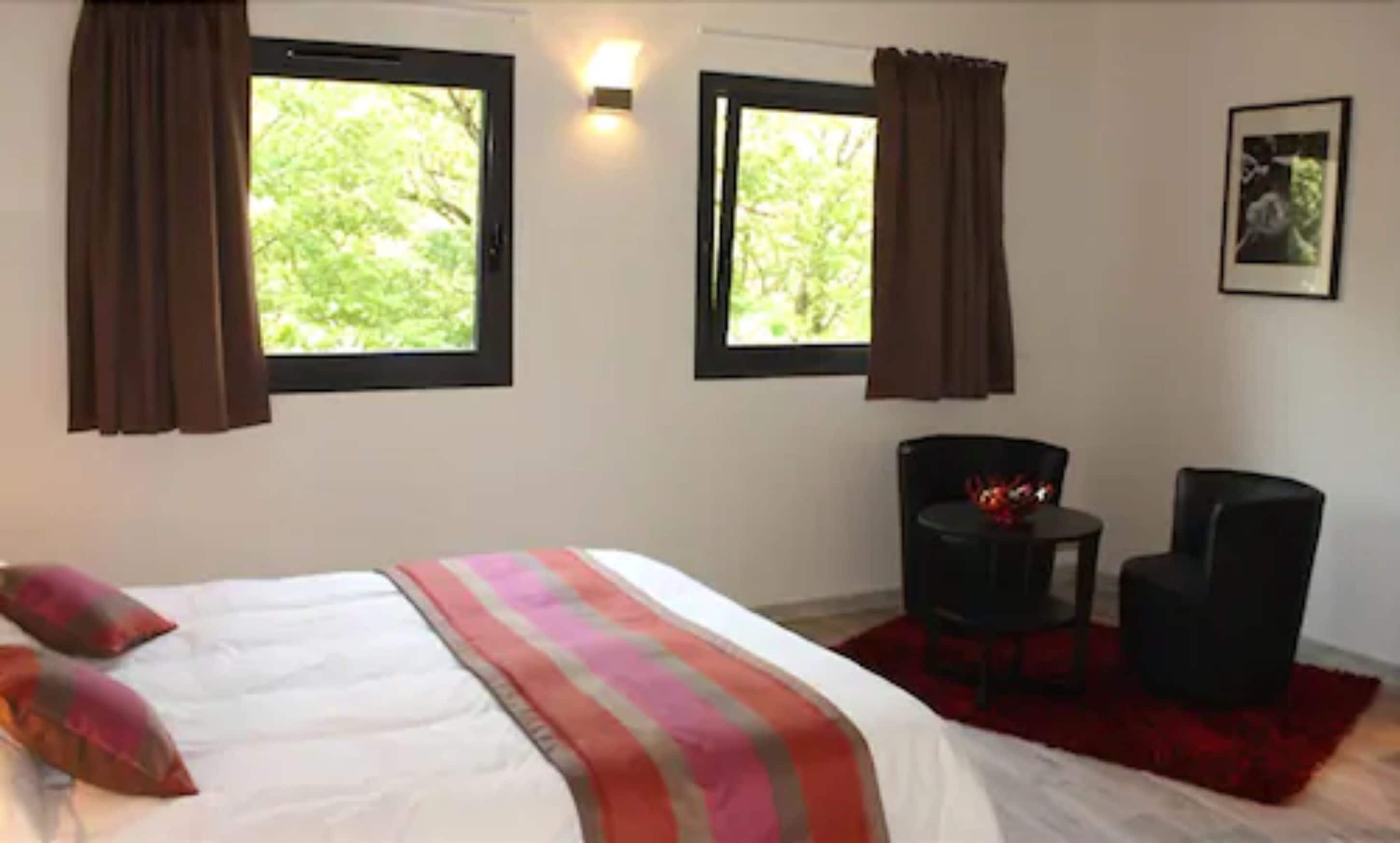 A bedroom with a bed, two windows with curtains, a chair, a small table, and a framed picture on the wall.