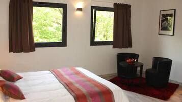 A bedroom with a bed, two windows with curtains, a chair, a small table, and a framed picture on the wall.