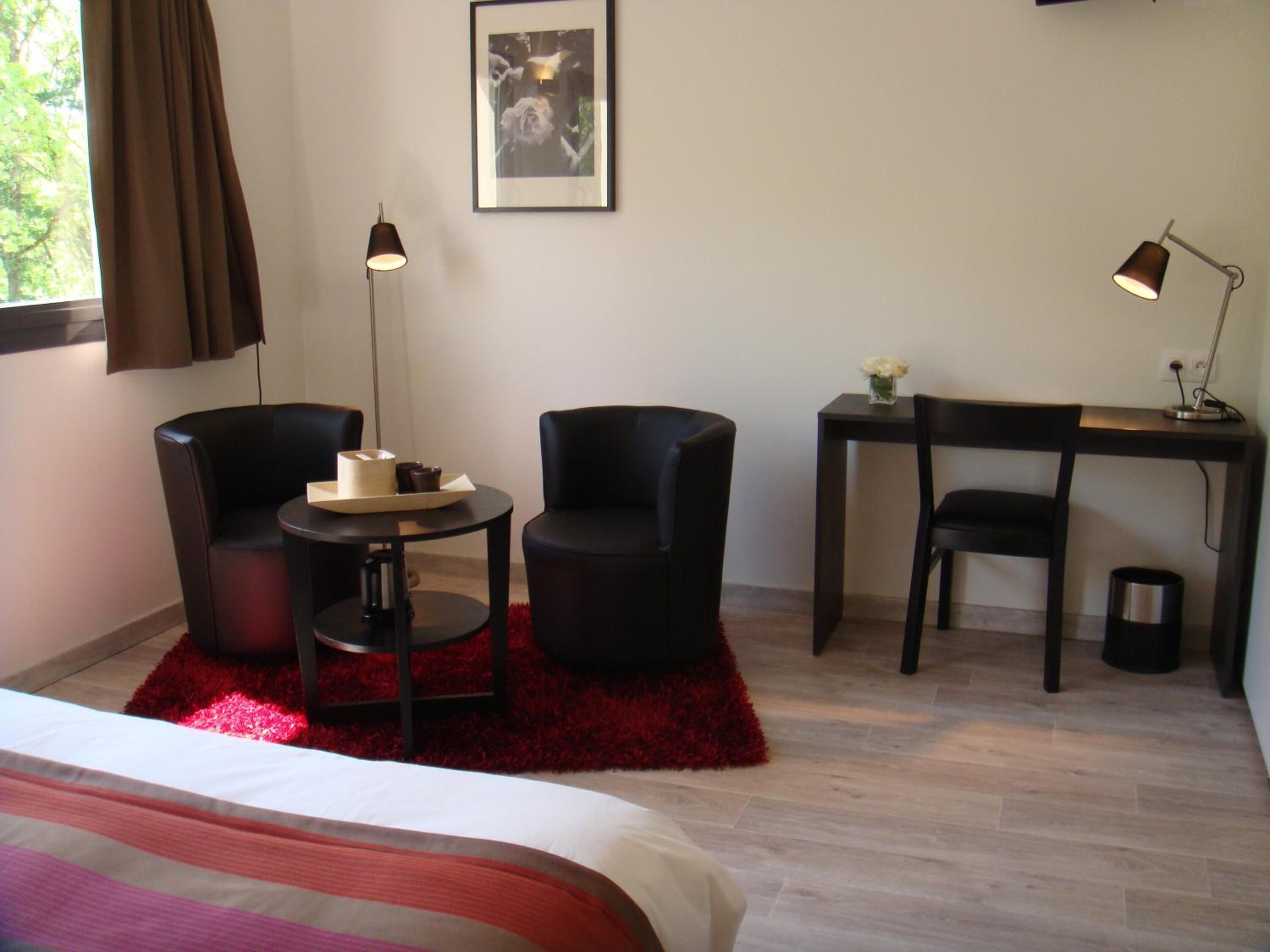 A hotel room with a bed, two black chairs, a small round table, a desk with a chair, a red rug, a framed picture on the wall, and two floor lamps.