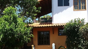 A white building with a tiled roof, a wooden door, and a small window. There is a paved pathway leading to the entrance, surrounded by various potted plants and greenery. - Pousada Barcarola (Búzios)