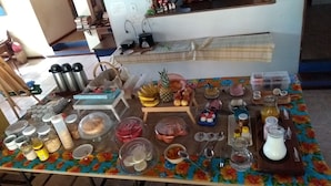 A table with various food items and kitchen utensils, a shelf with books and a computer, and a chair with a bag. - Pousada Barcarola (Búzios)