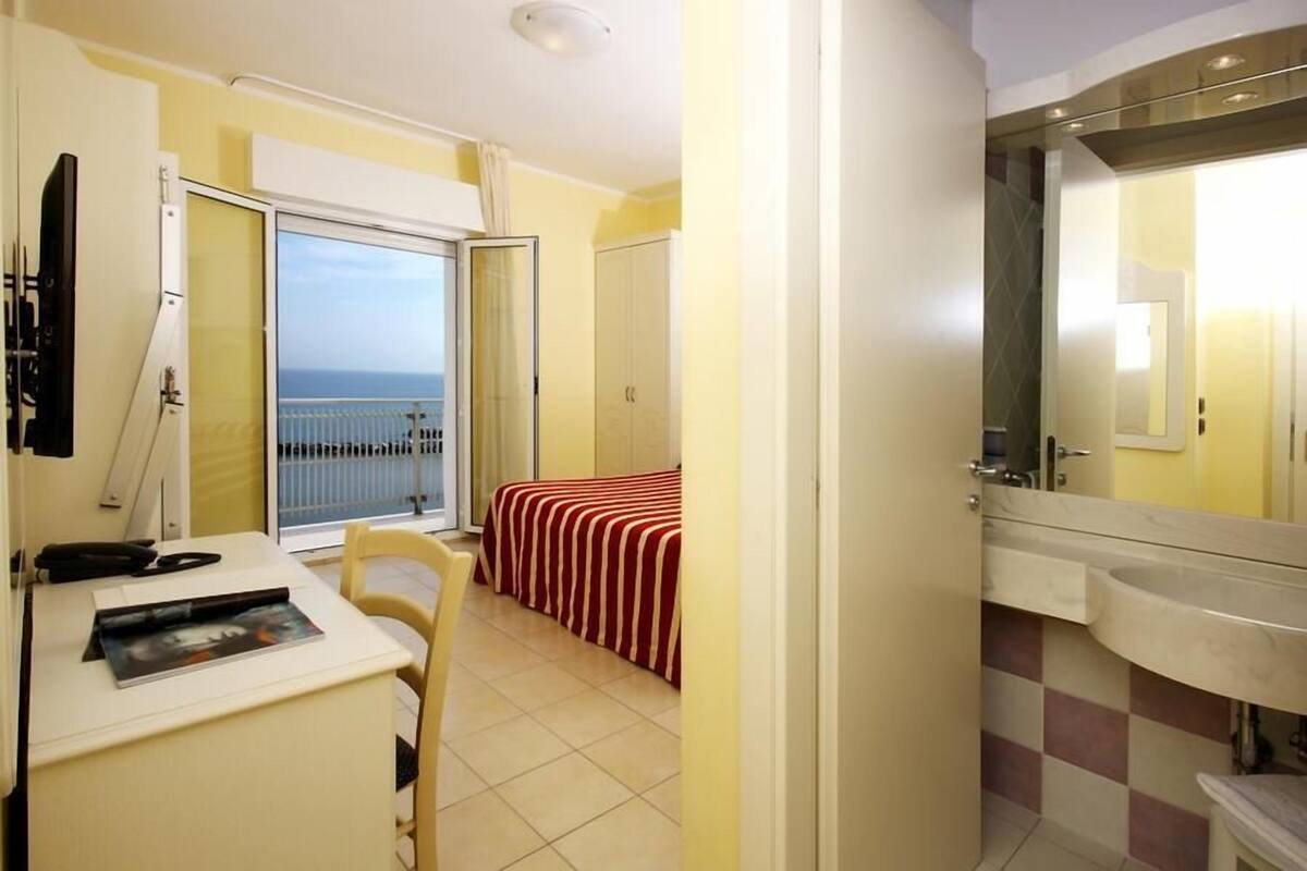 Una habitación de hotel compacta con balcón, cama con ropa de cama a rayas, escritorio pequeño con silla y baño con espejo grande y lavamanos.