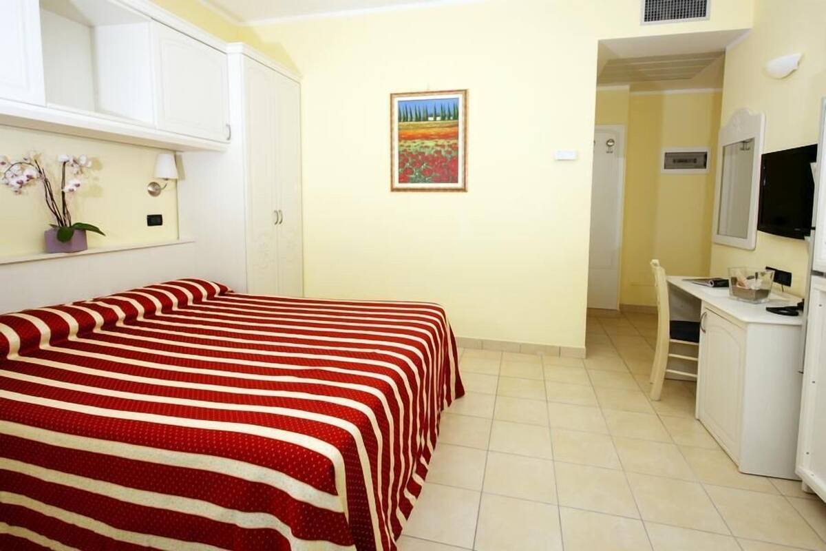 Un dormitorio con cama, escritorio, silla, televisor y un cuadro en la pared.