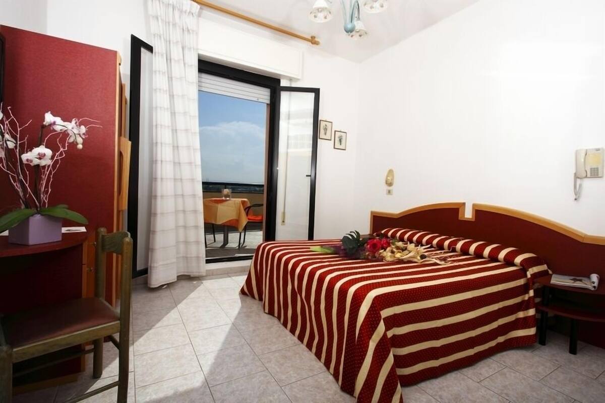 Habitación de hotel con cama, una silla, una mesa y vista al mar a través de una puerta abierta.