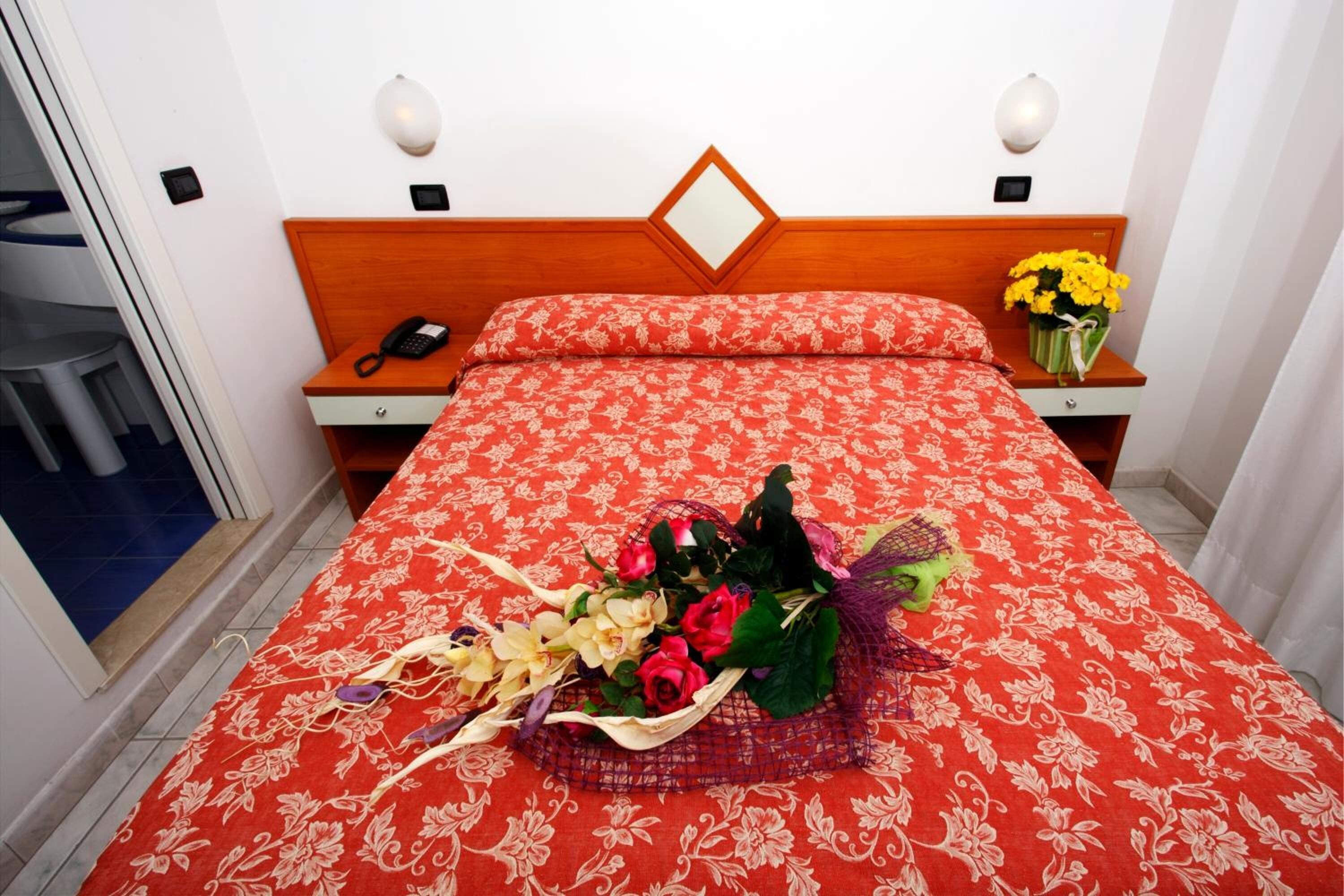 A hotel room with a wooden headboard, a red floral bedspread, a telephone on the nightstand, and a bouquet of flowers on the bed.
