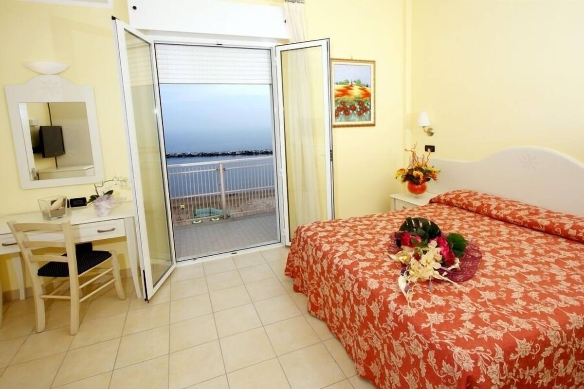 Habitación de hotel con balcón, una cama decorada con motivos florales, un escritorio con una silla y un cuadro en la pared.