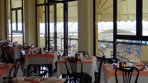 A restaurant with multiple tables and chairs, large windows with a view of the beach, and a tent-like structure outside. - Baia (Rimini)