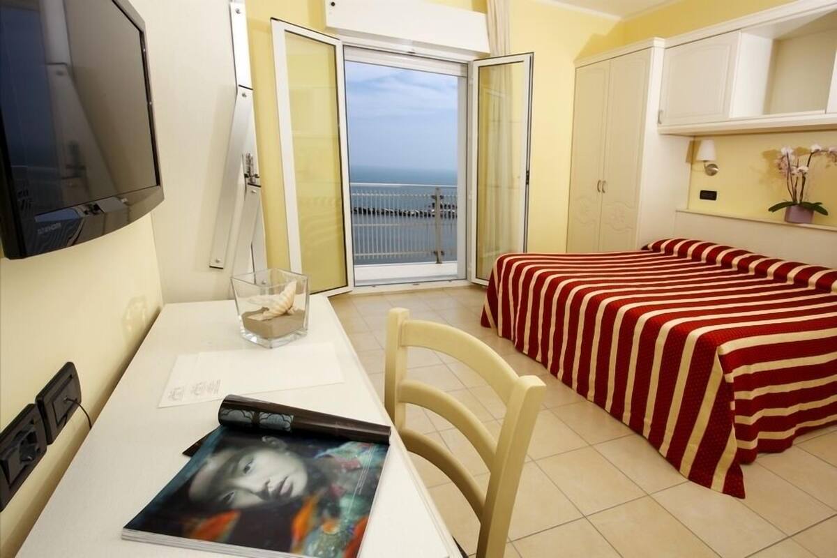Habitación de hotel con cama, una mesa con una revista, televisión y balcón con vista al mar.