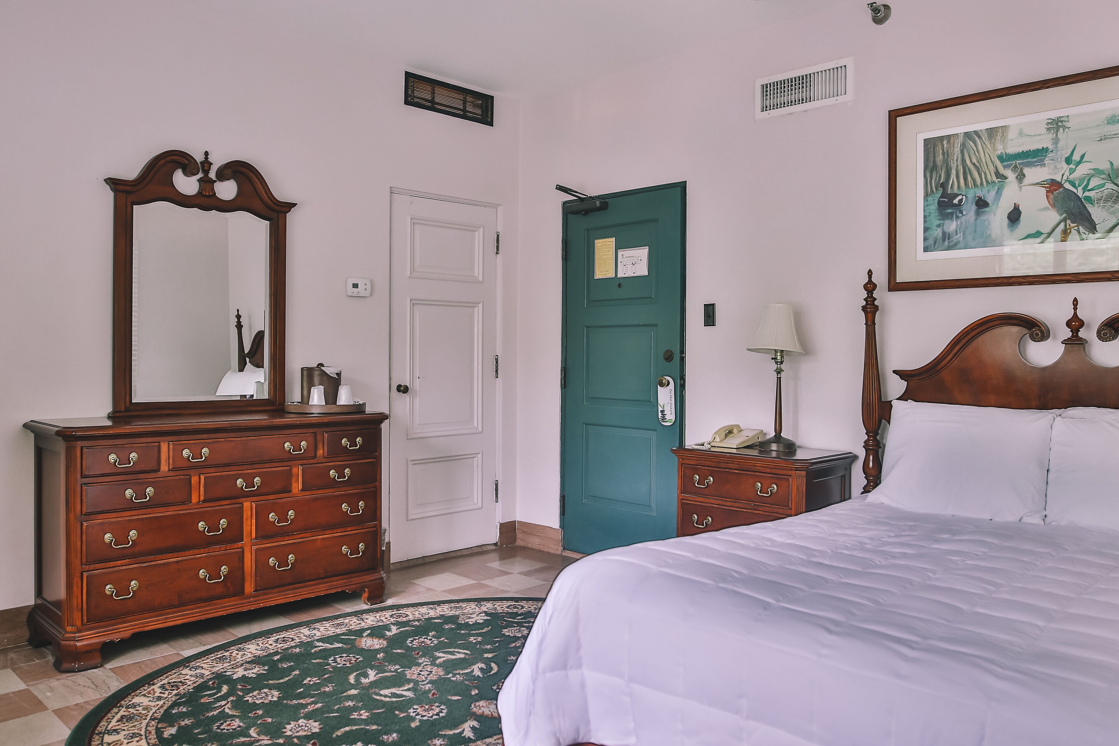 A bedroom with a large bed, a dresser, a green door, a framed picture on the wall, and a patterned rug.