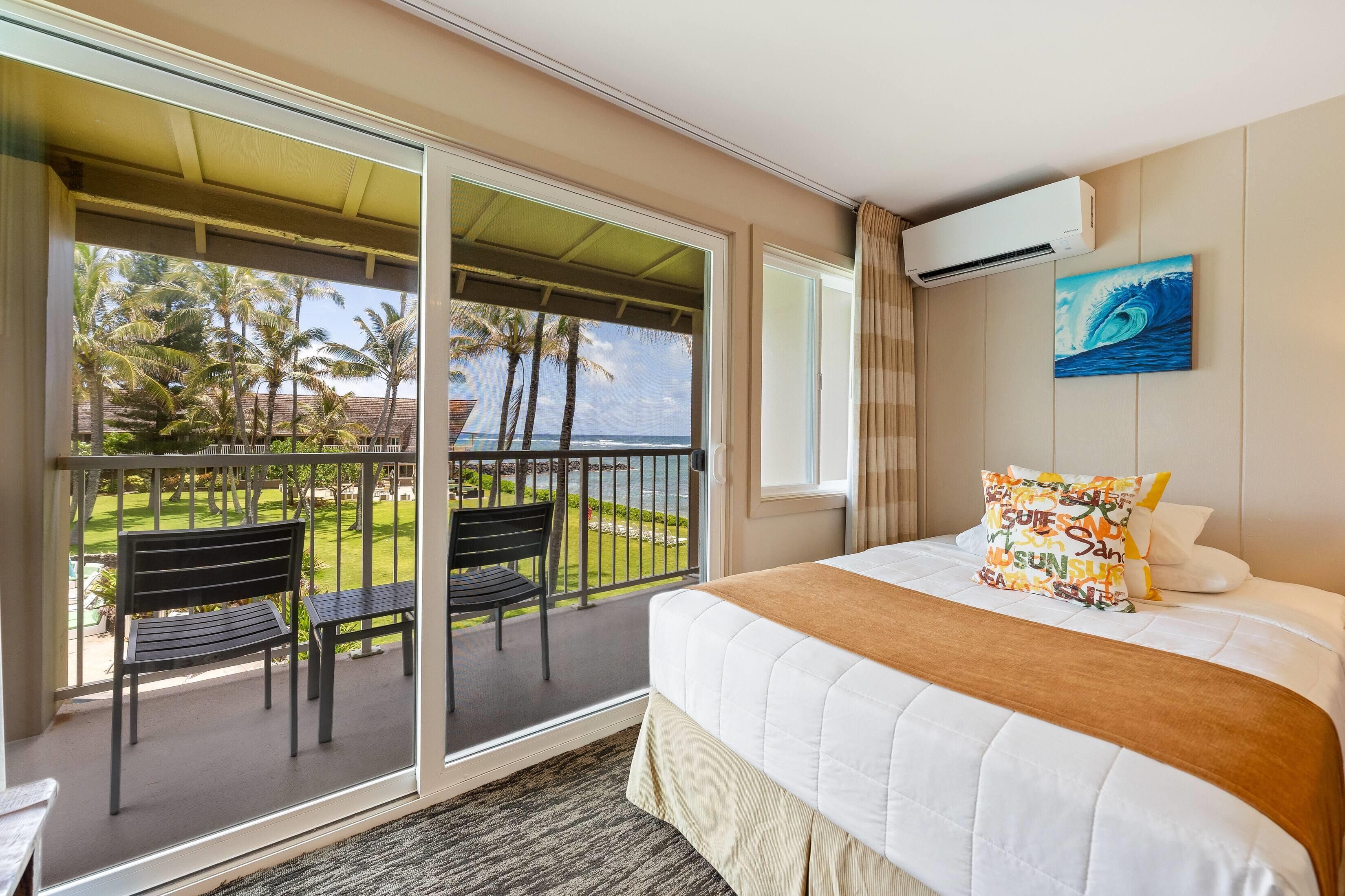 A hotel room with a balcony, a bed with a white comforter, and a painting of a wave.