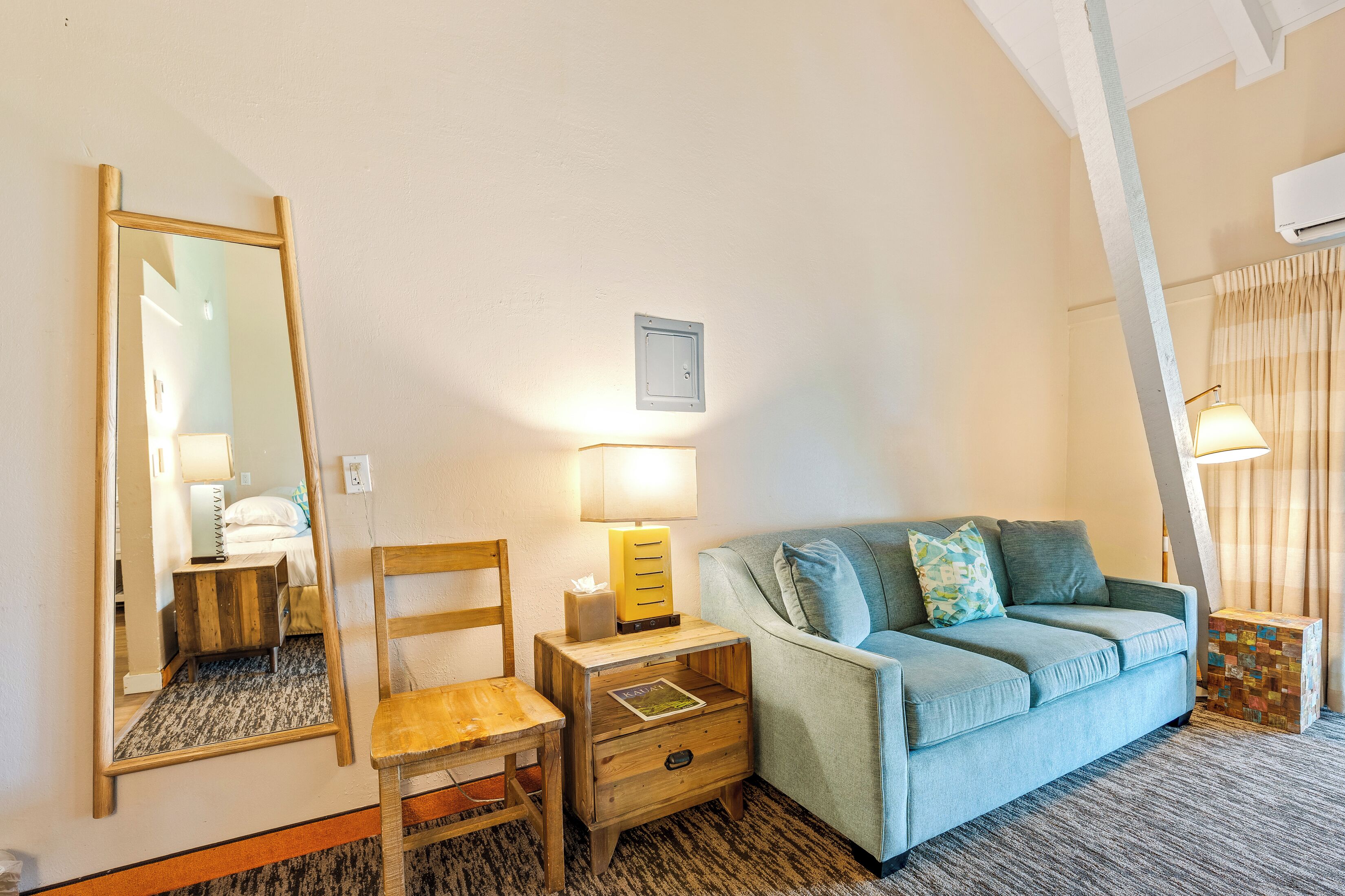 A room with a wooden chair, a small wooden nightstand with a lamp, a light blue sofa, and a mirror on the wall.