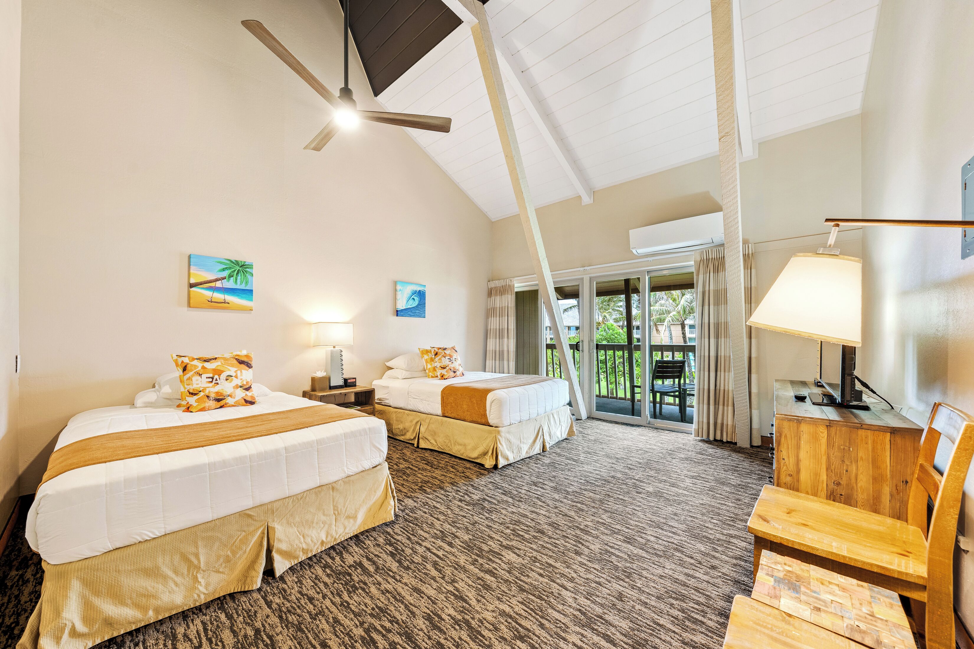 A hotel room with two beds, a desk, a chair, a ceiling fan, and a view of the outdoors.