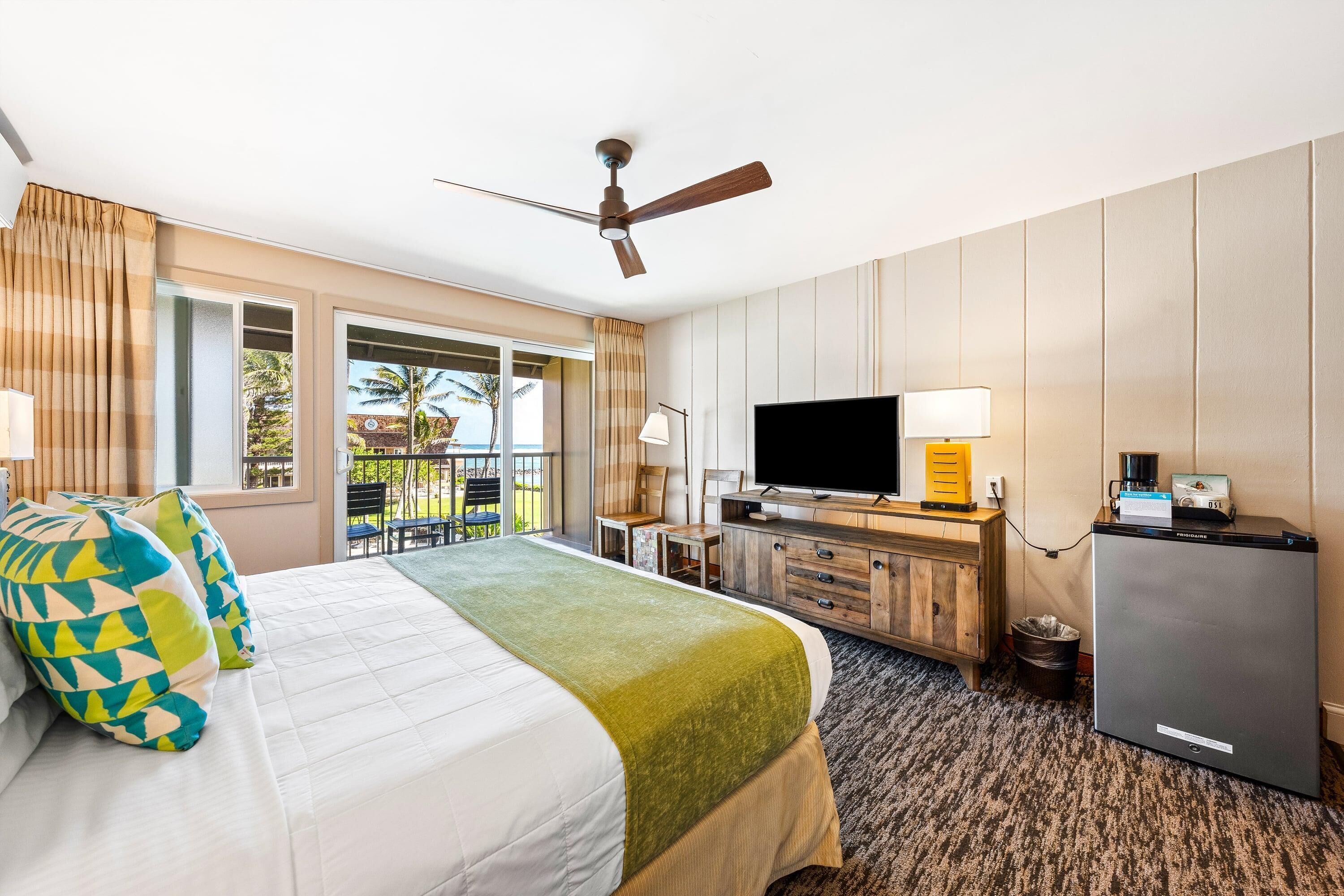 Habitación de hotel con cama, televisión, escritorio con ordenador y balcón con vistas a palmeras.